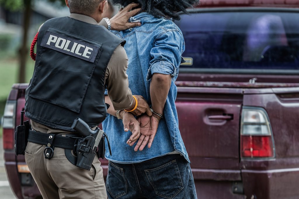 Police officer arresting man in handcuffs behind his back walking towards a red pickup truck